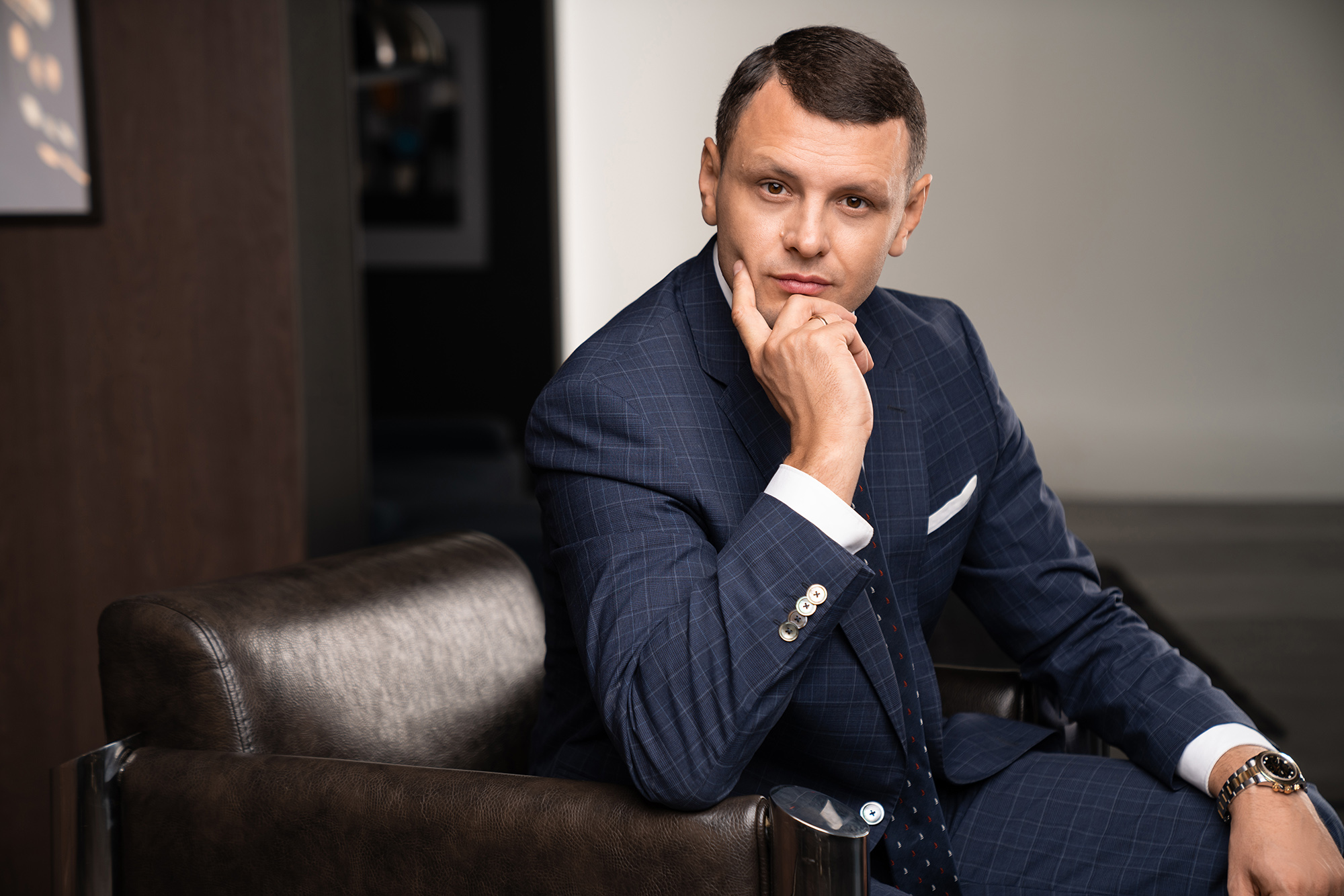 Чоловік у синьому піджаку сидить на коричневому кріслі і підтримує голову рукою