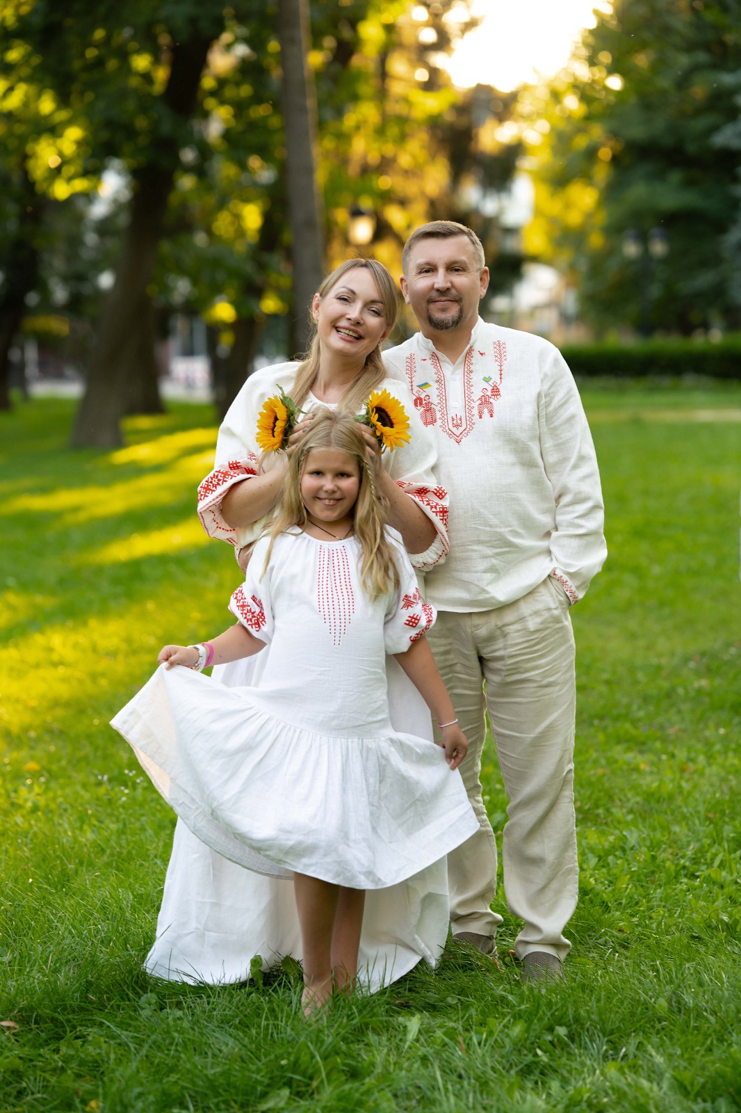 Мама, тато та донька у вишиванках посміхаються у літньому парку.
