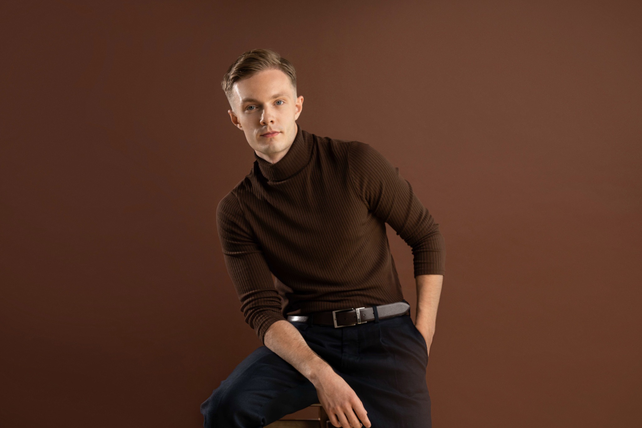 Молодий чоловік у коричневий светр сидить на стільці біля однотонного фону