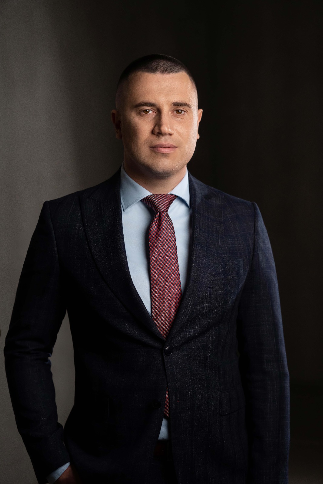 Чоловік у костюмі з червоною краваткою постановки на фоні сірої стіни.