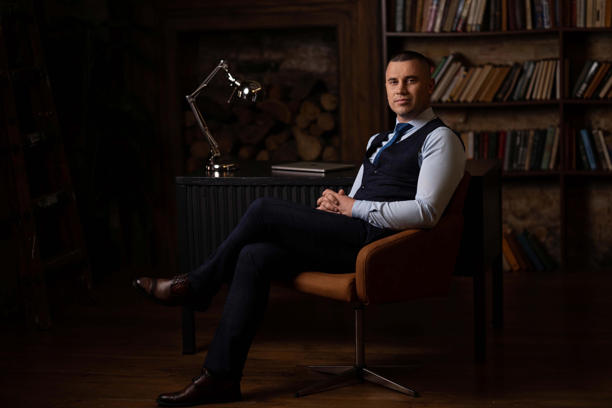 Чоловік у темно-синьому костюмі з жилетом сидить за робочим столом біля книжкових полиць, втілюючи атмосферу кабінету адвоката або партнера-керуючого.